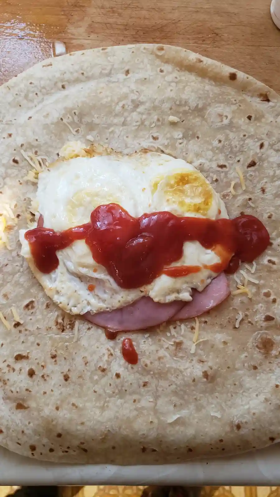 A close-up photo of a breakfast burrito in progress, showing a flour tortilla with a fried egg, ham, shredded cheese, and a…