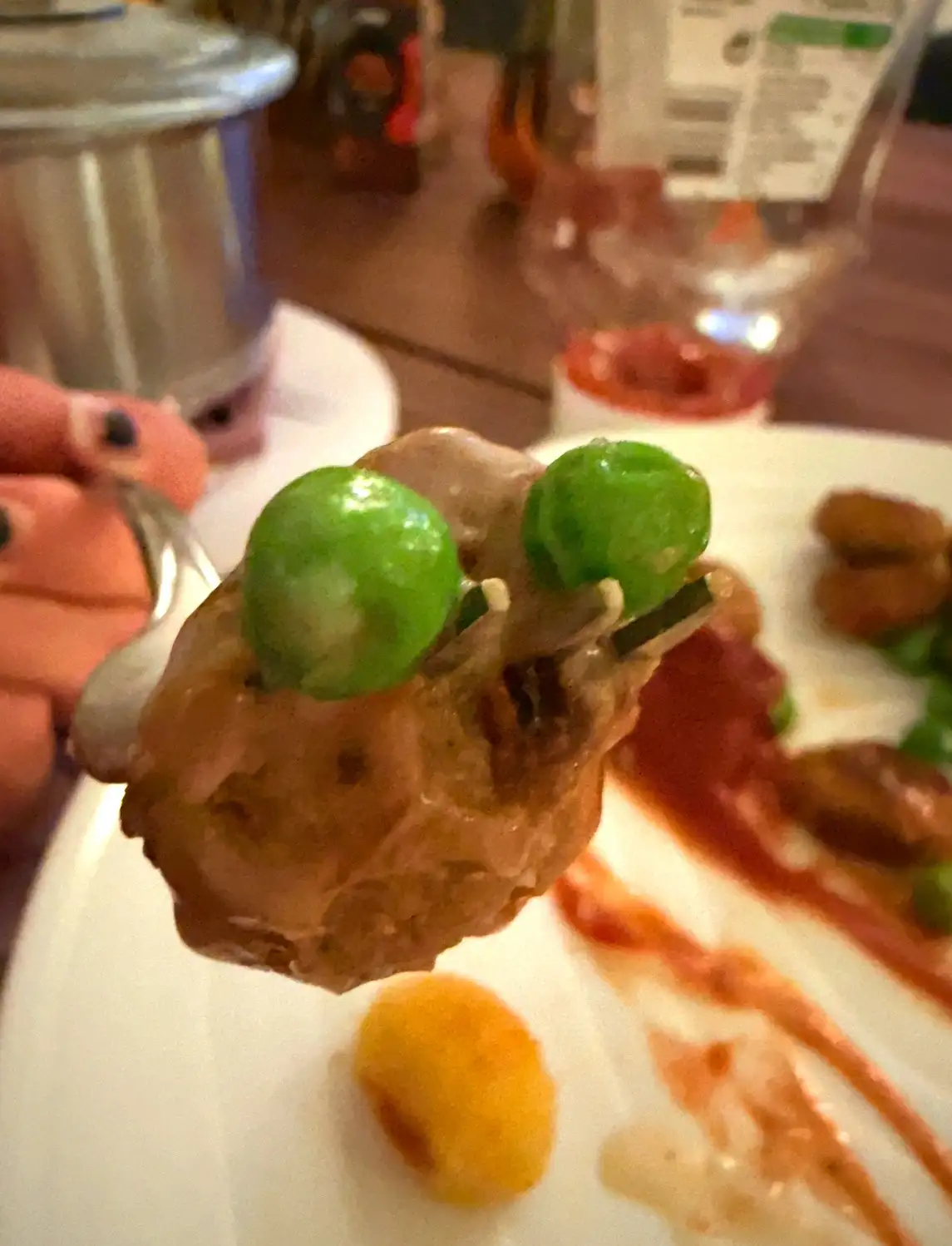 A close-up photo shows a fork holding a piece of food, possibly a dumpling or meatball, topped with two bright green peas…