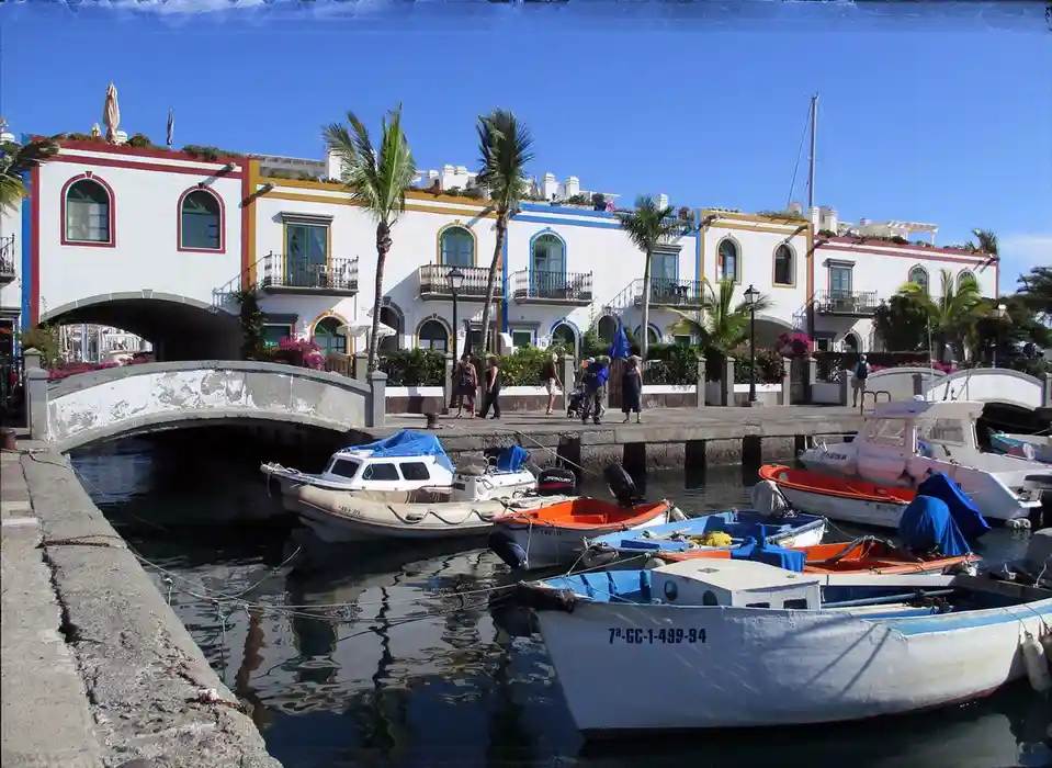 A scenic photo of a canal with colorful boats docked, featuring white buildings with arched windows and palm trees under a…