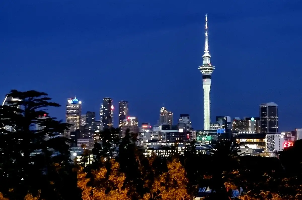 A nighttime cityscape featuring the illuminated Sky Tower in Auckland, New Zealand, rising above surrounding skyscrapers…