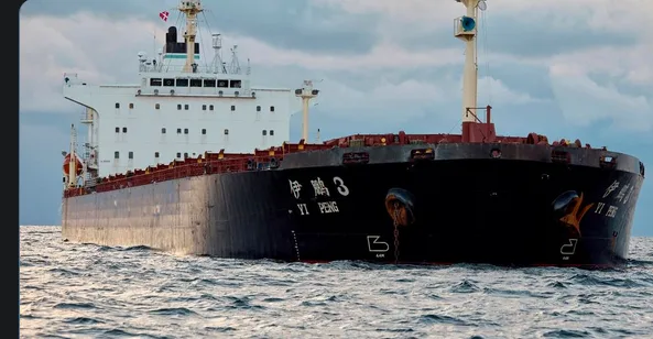 A large black cargo ship named '伊鹏3' (Yi Peng 3) sails on choppy ocean waters under a cloudy sky. The ship's name is written…