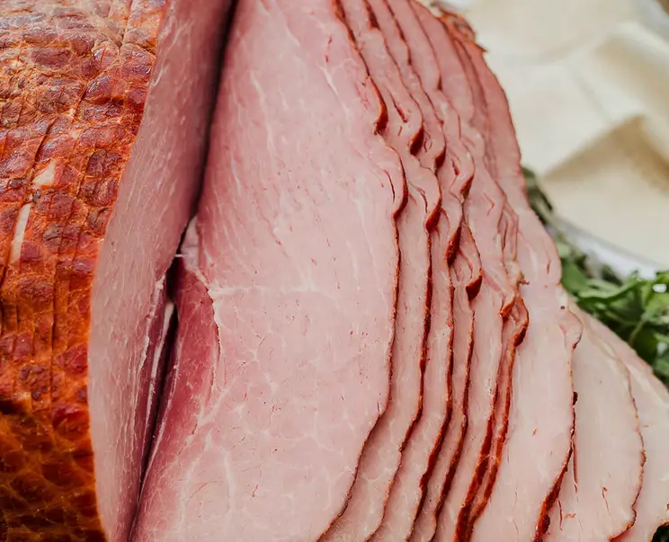A close-up photo of a large, roasted ham with a crispy, browned exterior and numerous thin, pink slices cut from its side…