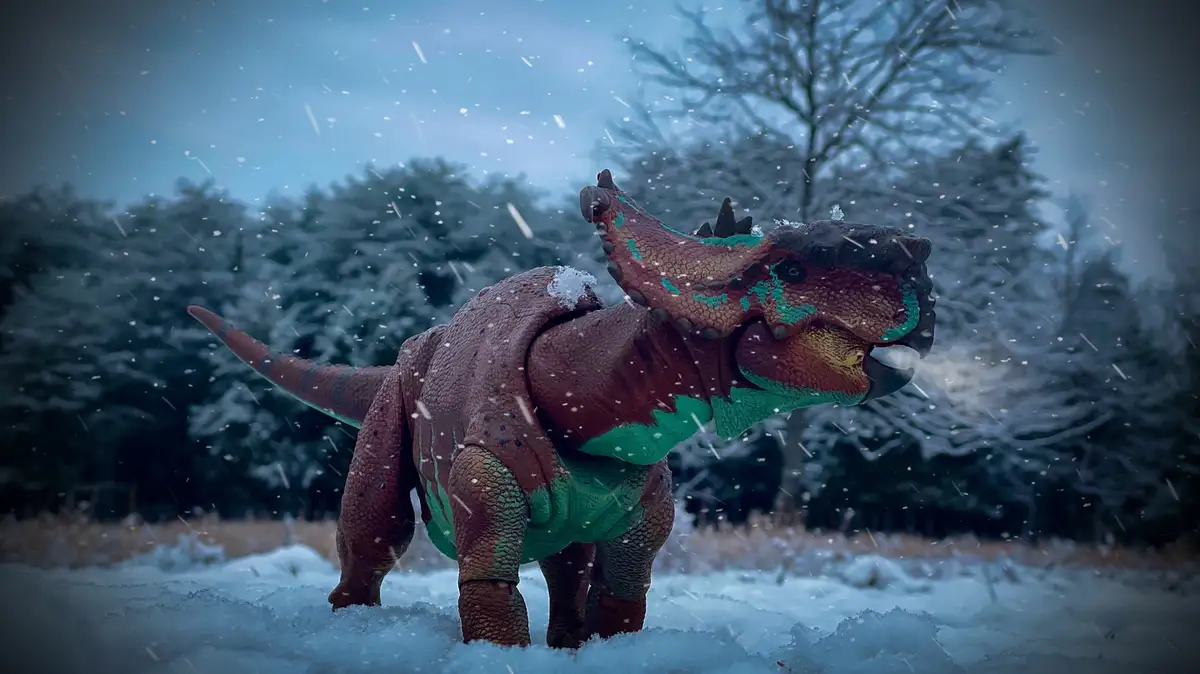 A toy dinosaur figurine, resembling a ceratopsian with brown and green coloring, stands in a snowy outdoor scene with snow…