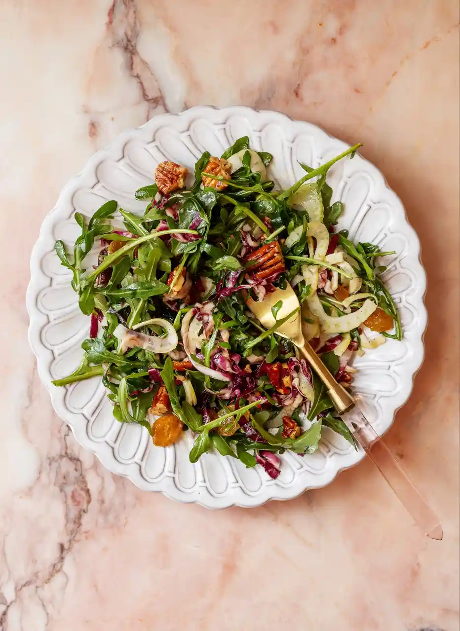 An overhead shot of a colorful salad in a white, scalloped ceramic bowl on a pink marble surface. The salad contains…