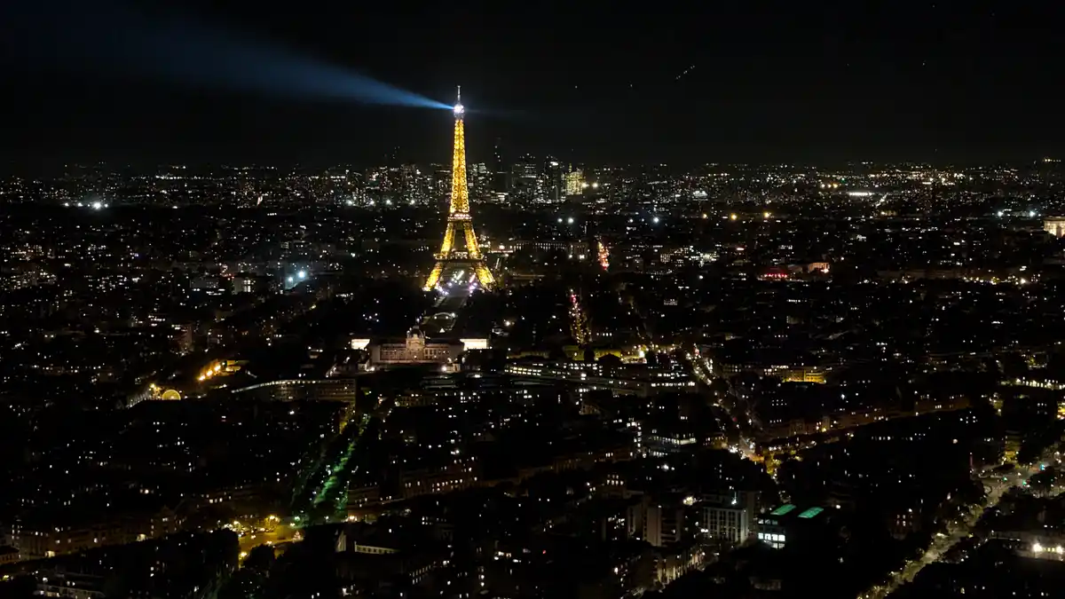 A nighttime aerial view of Paris, with the Eiffel Tower illuminated in gold and projecting a blue light beam into the sky…