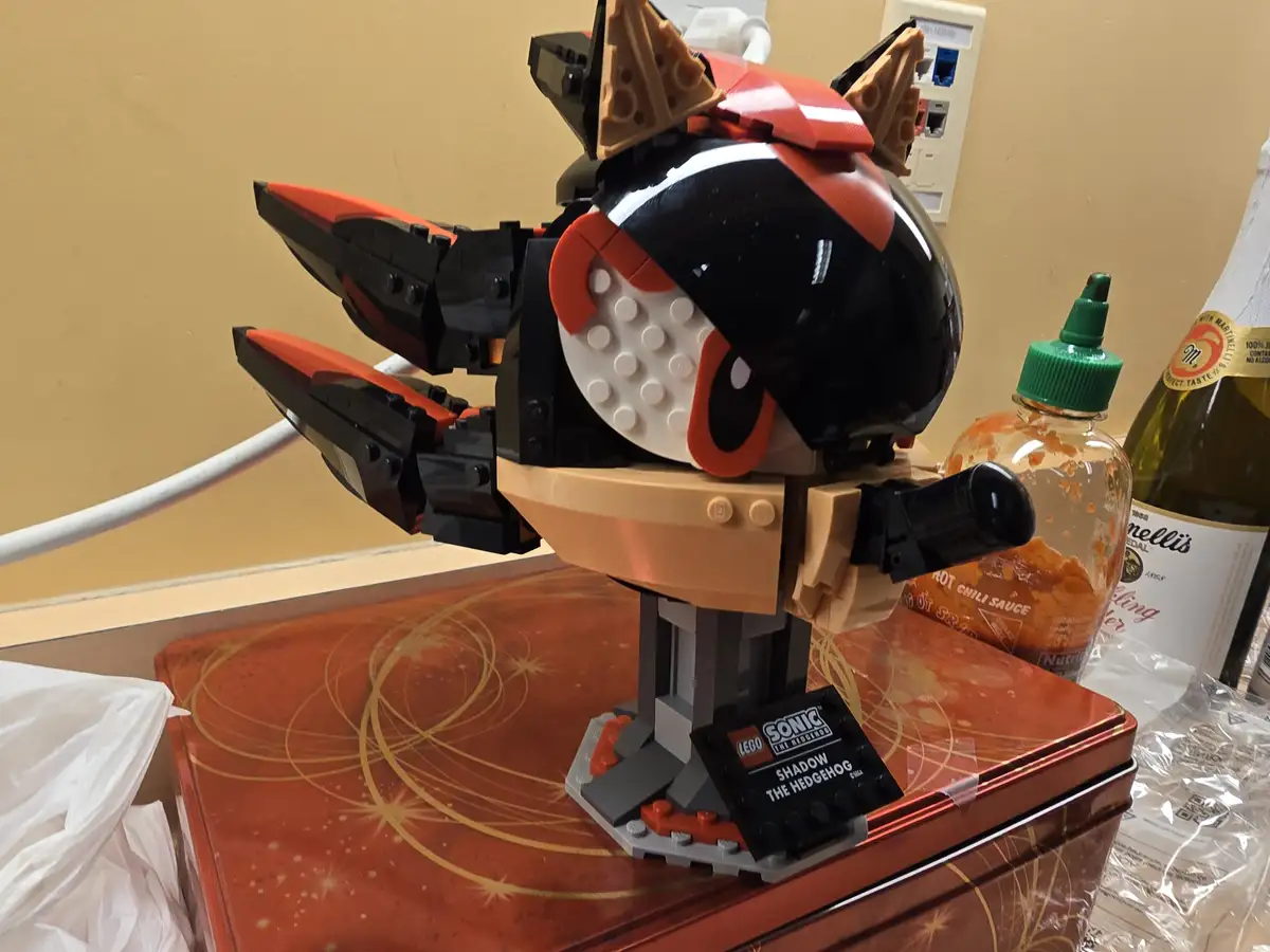 A close-up of a LEGO Shadow the Hedgehog minifigure head, mounted on a stand and placed atop a decorative red tin. The…