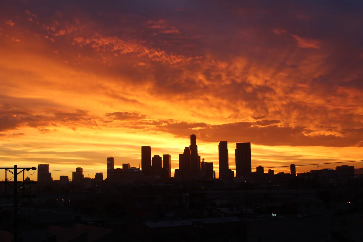 A dramatic sunset over a city skyline, with fiery orange and red clouds above dark silhouetted skyscrapers and foreground…