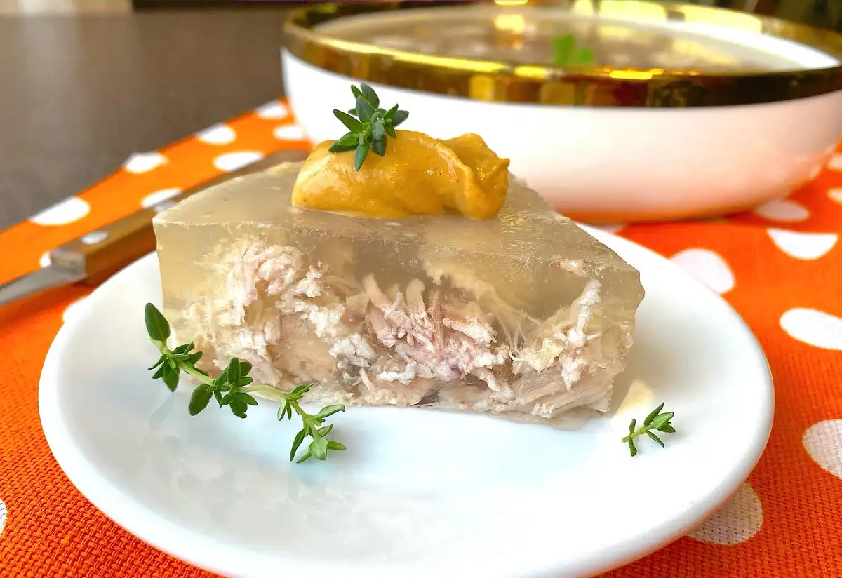 A slice of translucent meat jelly, containing shredded meat inside, is presented on a white plate with a dollop of orange…