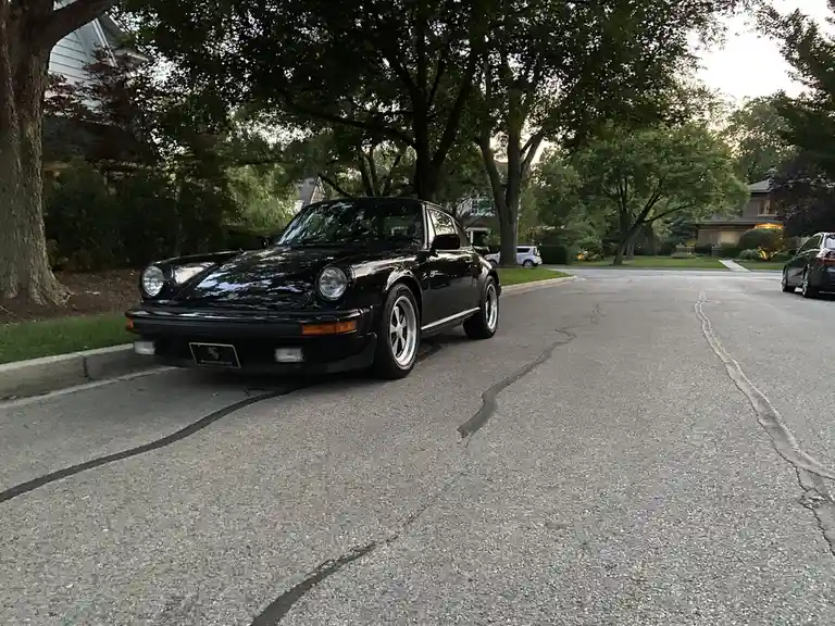A black Porsche 911 is parked on a residential street lined with trees and houses, with the overlaid text reading 'this is a…