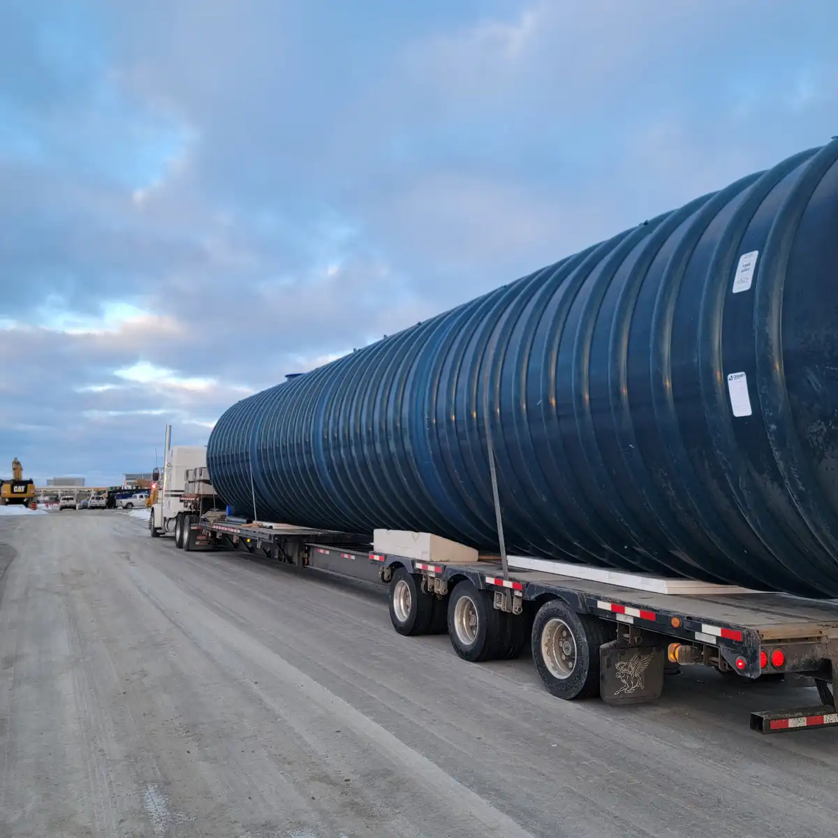 A large, dark blue corrugated cylindrical tank is being transported horizontally on a long flatbed trailer pulled by a white…