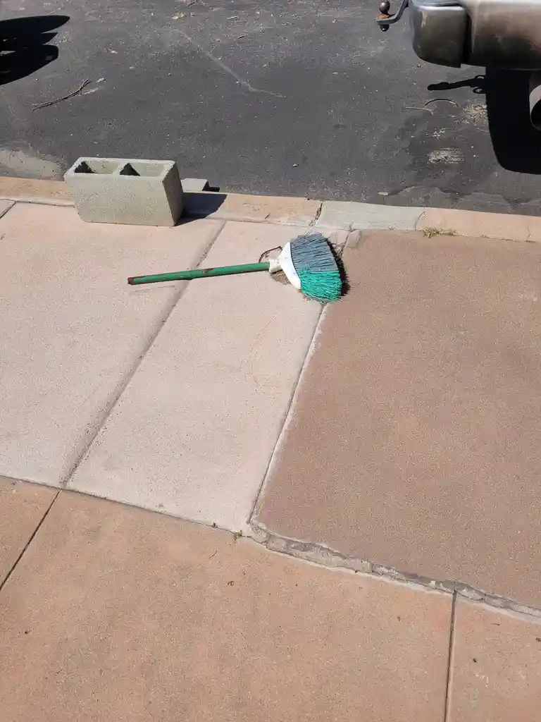 A green-handled broom with gray and teal bristles lies on a sidewalk next to a concrete block, with a vehicle's bumper…