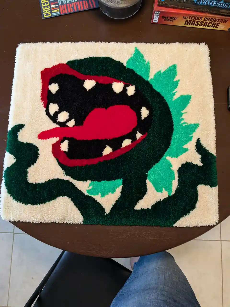 A fluffy, beige rug with a large, cartoonish green monster wearing a red Santa hat, featuring a wide open mouth with red…
