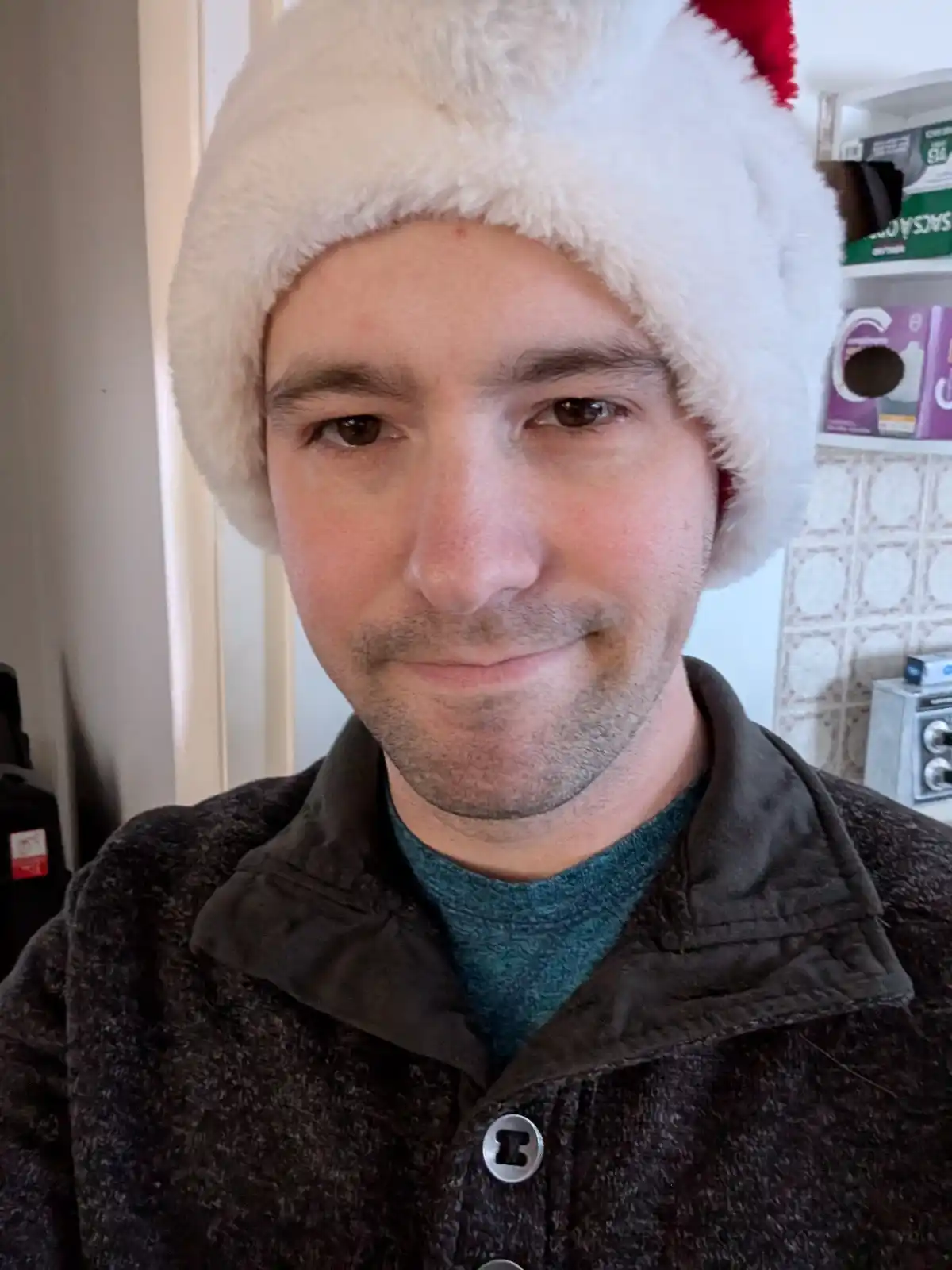 A man wearing a white and red Santa hat with a subtle, unenthusiastic smile looks at the camera. He wears a dark textured…