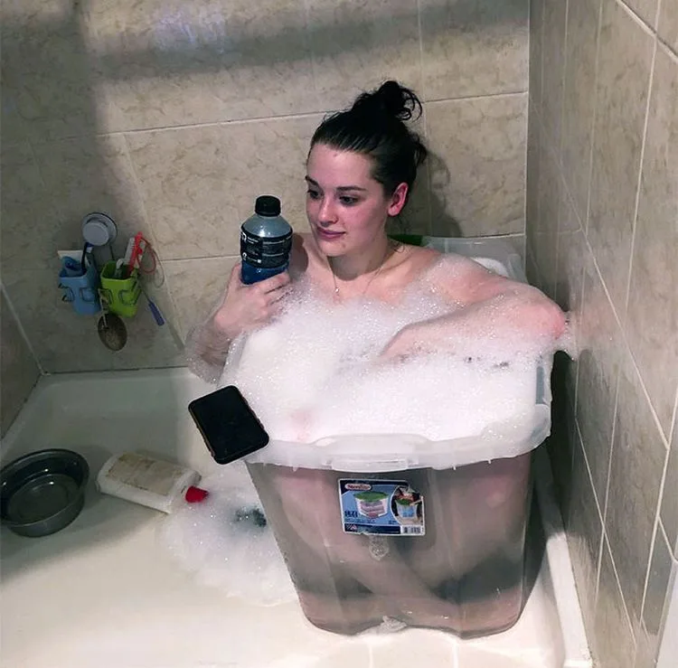 A young woman with dark hair in a bun sits in a bathtub filled with bubbles, submerged in a large clear plastic storage bin…
