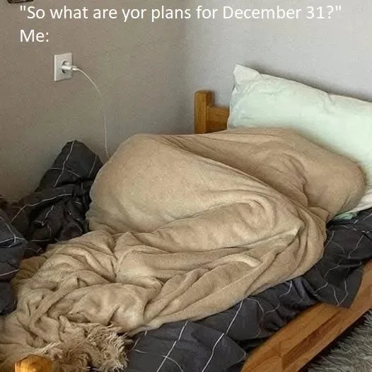 A person is buried under a tan blanket in a messy bed, with only their head visible, while a white charging cable plugs into…