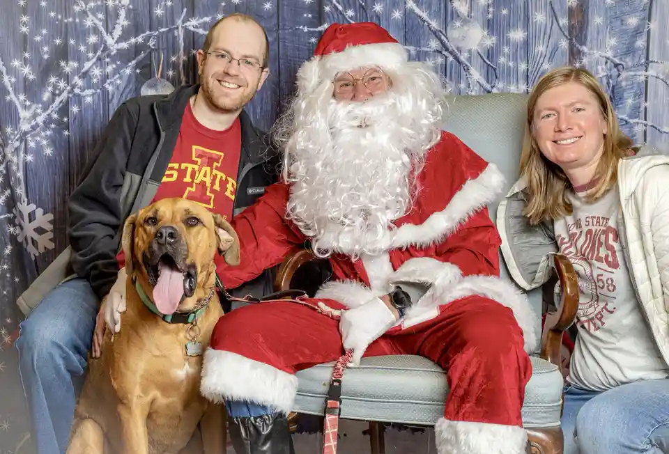 A man, woman, and their brown dog pose with a person dressed as Santa Claus in a holiday photo booth. The Santa figure…