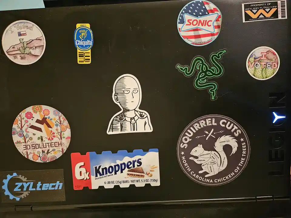 A black laptop lid covered in various stickers including 'I Voted' Texas and cactus designs, a Chiquita banana logo, a Sonic…