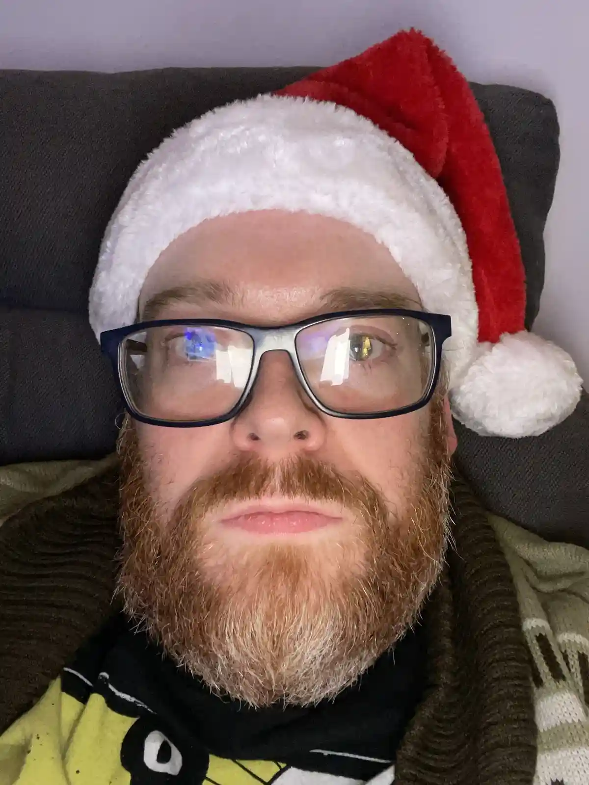 A man with a reddish beard and glasses wears a red and white Santa hat while lying on a dark couch. He has a neutral…