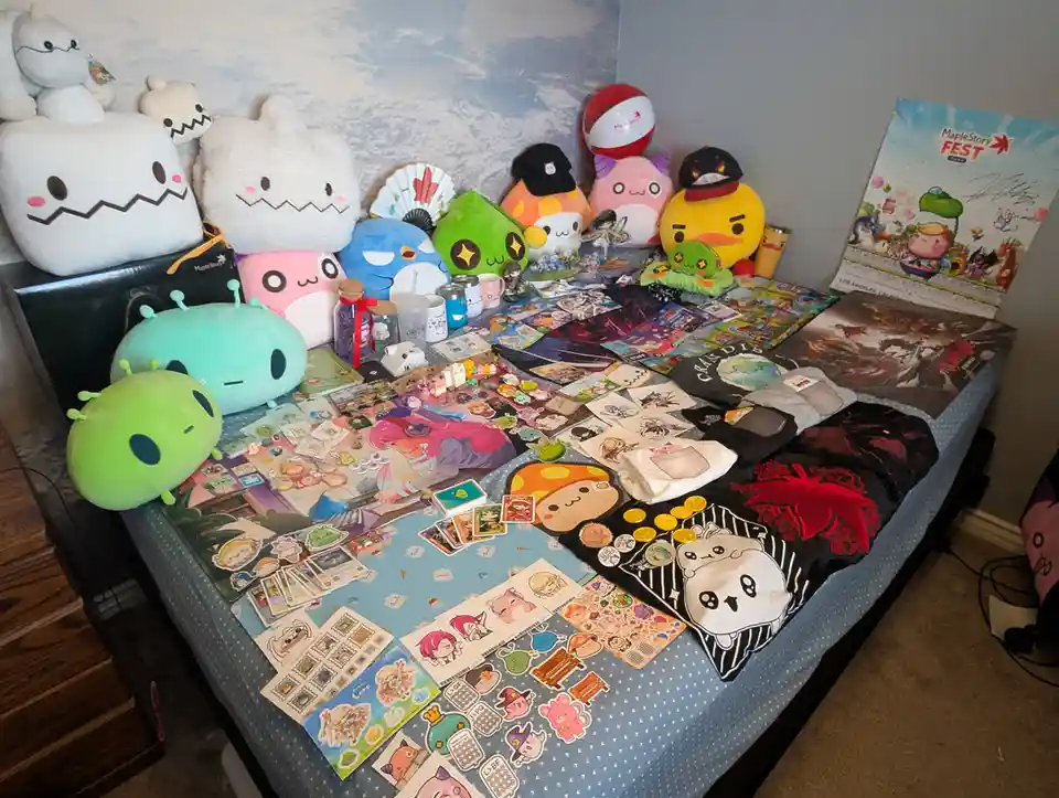 A bed is covered in a massive collection of MapleStory merchandise, including plushies, t-shirts, stickers, cards, and a…