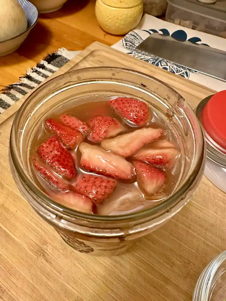A glass jar filled with sliced strawberries submerged in a clear liquid sits on a wooden cutting board. The jar is on a…