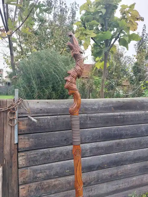 A highly ornate, carved wooden staff with a dragon-like head and swirling patterns leans against a weathered wooden fence in…
