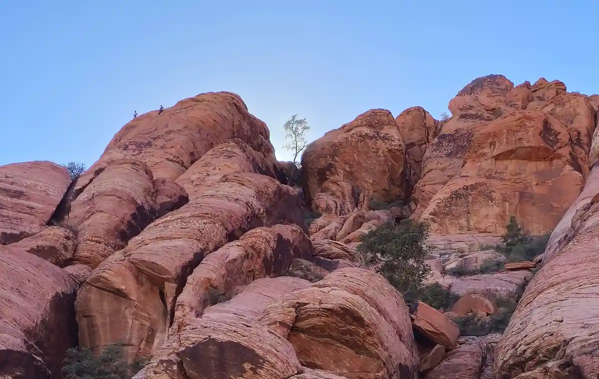 A wide-angle photo shows two tiny human figures standing atop large, rounded red rock formations under a clear blue sky…