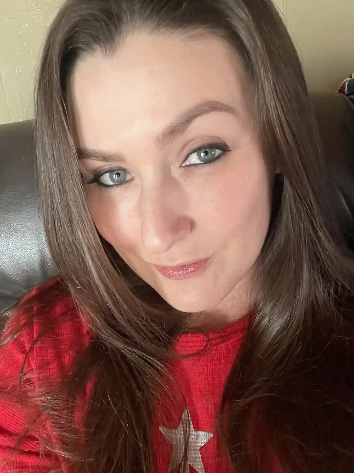 A close-up selfie of a woman with long brown hair and blue eyes, wearing a red sweater with a silver star, looking directly…