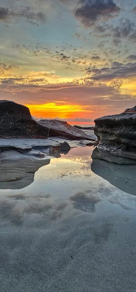 A serene sunset photo shows a calm tide pool reflecting the orange and yellow sky between large rocks. Overlaid text reads…