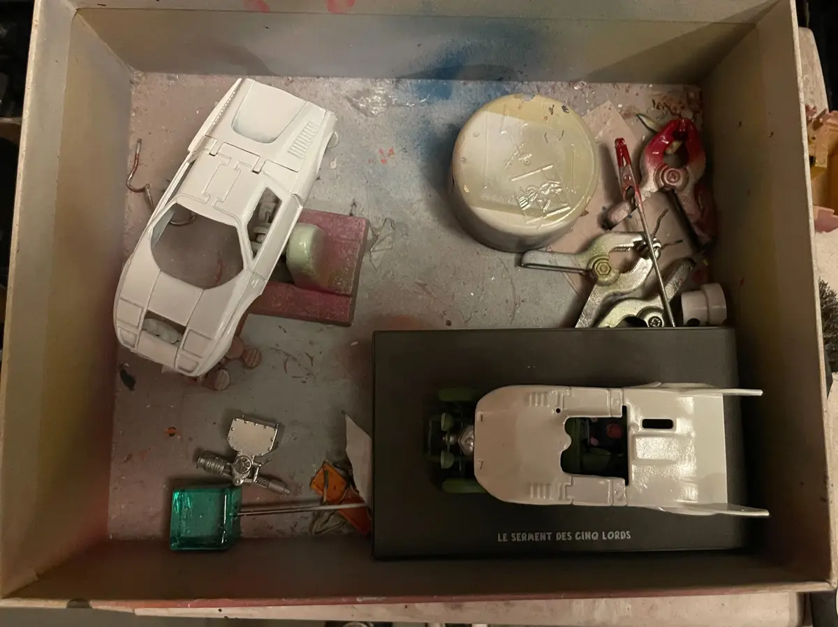 An overhead shot of a cardboard box containing a white model car on a stand, various tools, and a second model car in a…