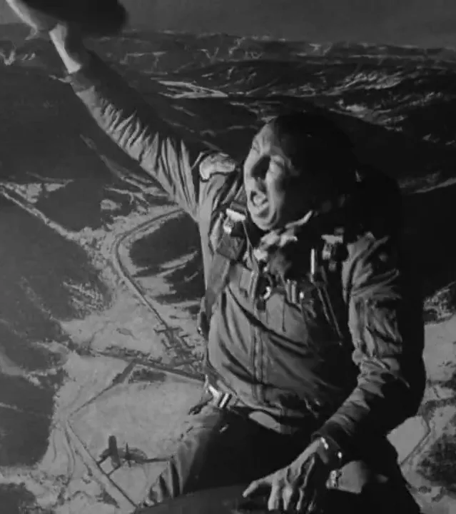 A black-and-white photo shows a man in a military-style jacket, seated and looking down at a valley below with his mouth…