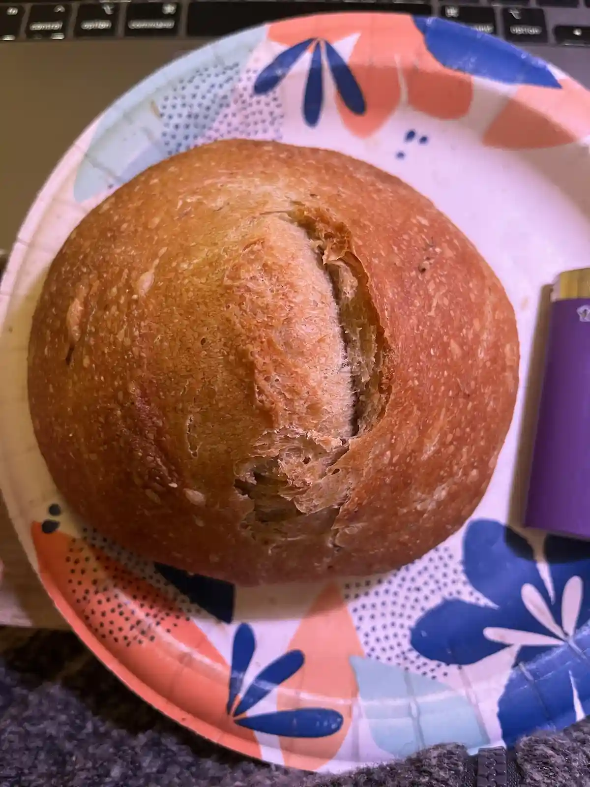 A round, golden-brown loaf of bread sits on a colorful paper plate with blue and orange floral patterns. A purple lighter…