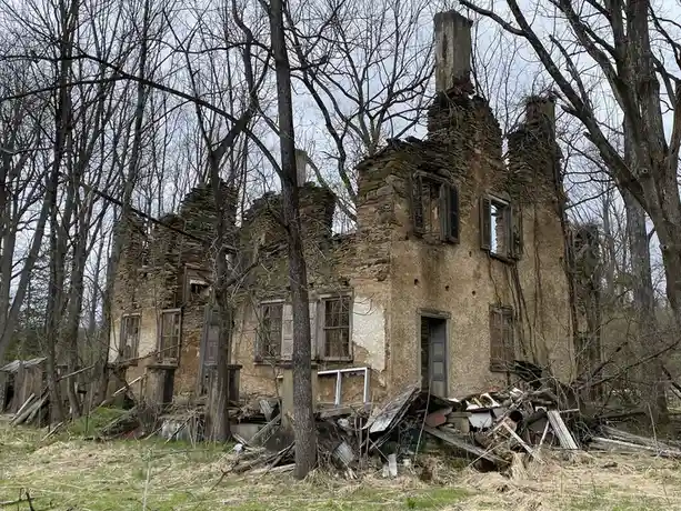 A dilapidated stone house with broken windows, collapsed roof sections, and rubble piled at its base, surrounded by bare…
