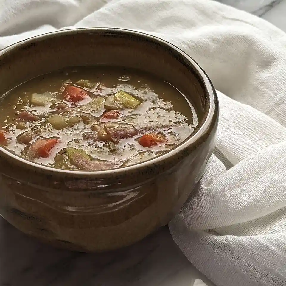 A close-up photo shows a rustic brown ceramic bowl filled with chunky vegetable soup, resting on a white cloth over a marble…