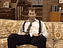 A man in a white shirt, black pants, and suspenders sits on a patterned couch in a living room. He slowly raises his right…