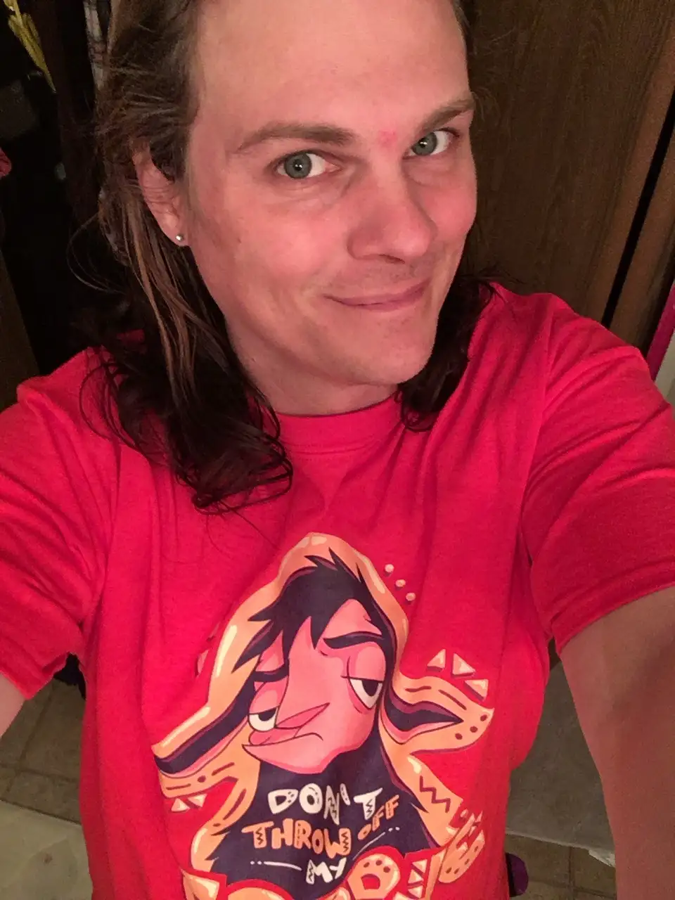 A person with long brown hair and a silver earring wears a red t-shirt featuring a cartoon character with a smug expression…