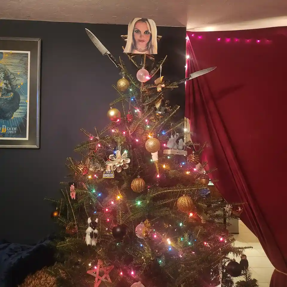 A decorated christmas tree with colorful lights and ornaments has a cutout of britney spears at the top, with two kitchen…