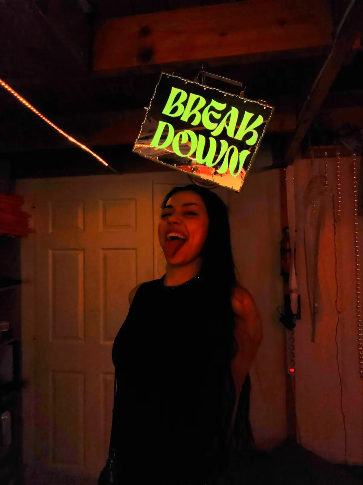 A person with long dark hair, wearing a black top, is laughing with their tongue out in a dimly lit room. Suspended directly…