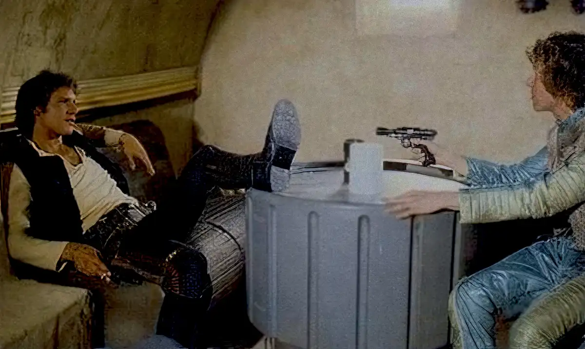 A man sits relaxed in a chair with his feet up on a table, while another person across the table points a blaster at him…