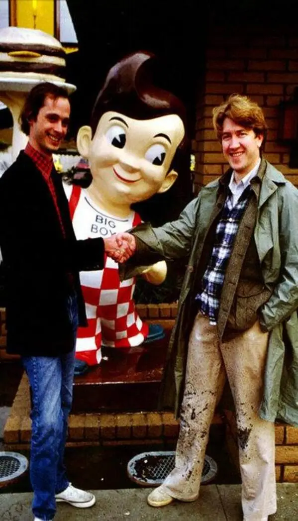 Two men stand beside a large, cartoonish statue of Big Boy, a vintage fast-food mascot wearing a red-and-white checkered…