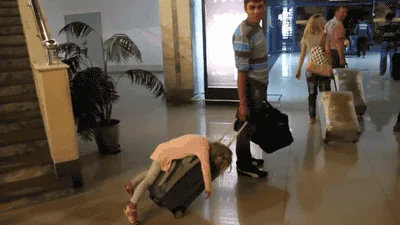 A sequence of images shows a woman lying horizontally on top of a rolling suitcase while a man walks beside her, seemingly…