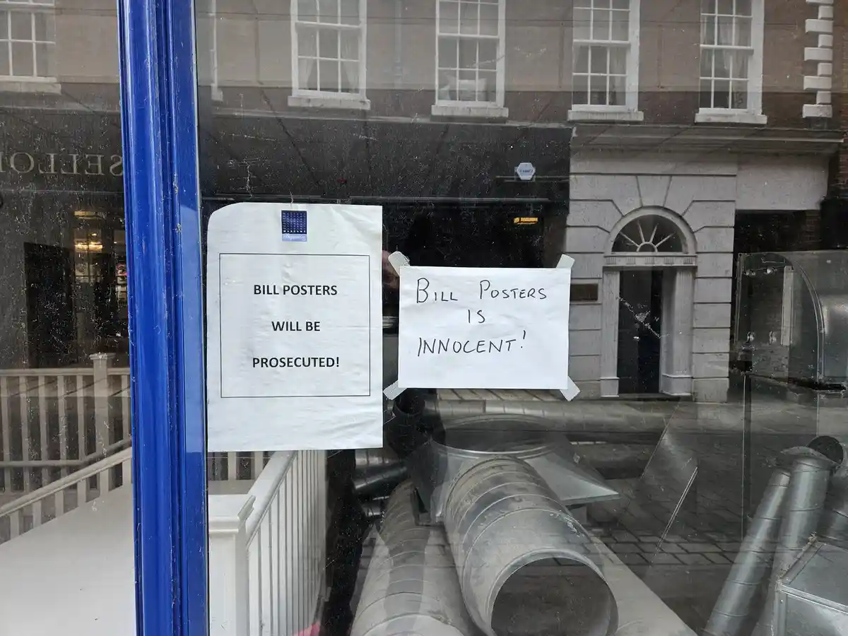 Two signs taped to a dirty storefront window: one official-looking printed sign says 'BILL POSTERS WILL BE PROSECUTED!' and…