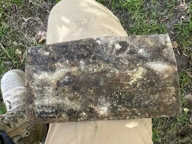 A person wearing beige pants and a muddy hiking boot holds a large, dirty, rectangular brick with black and brown stains and…
