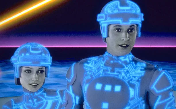 Two characters from the movie Tron, a man and a woman in glowing blue digital suits with helmets, stand against a neon-lit…