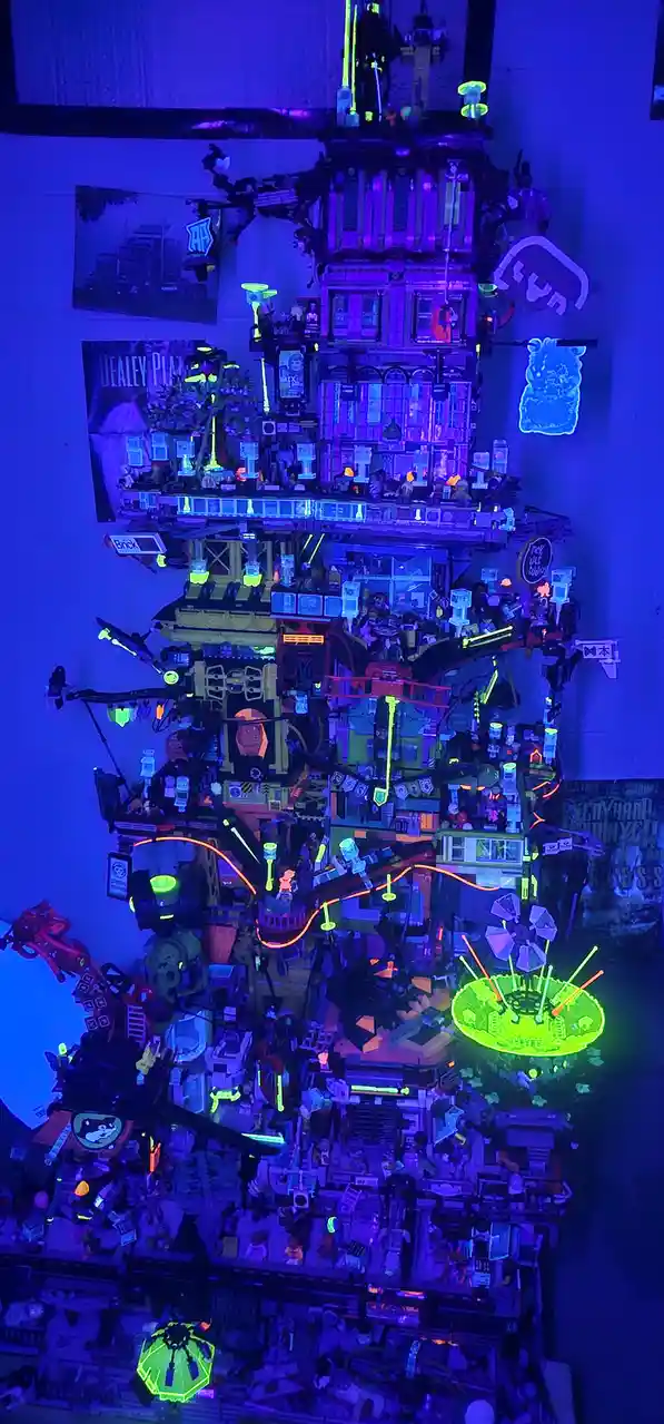 a towering, intricate lego structure illuminated under blacklight, glowing with vibrant neon purple, green, and orange…