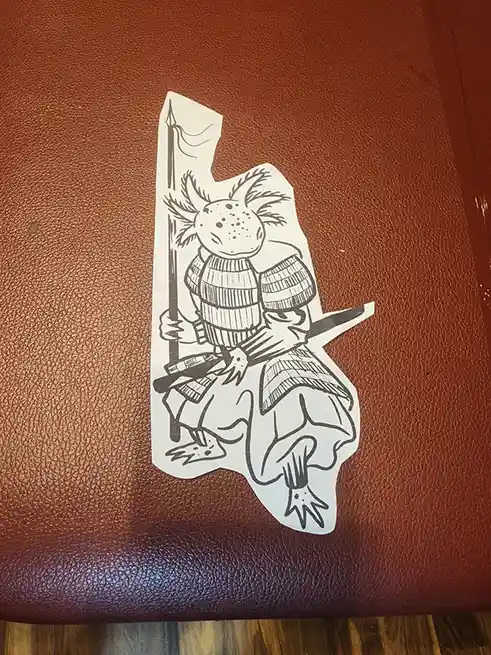 A black-and-white sticker depicts an axolotl wearing samurai armor, holding a spear and a katana, sitting on a brown…