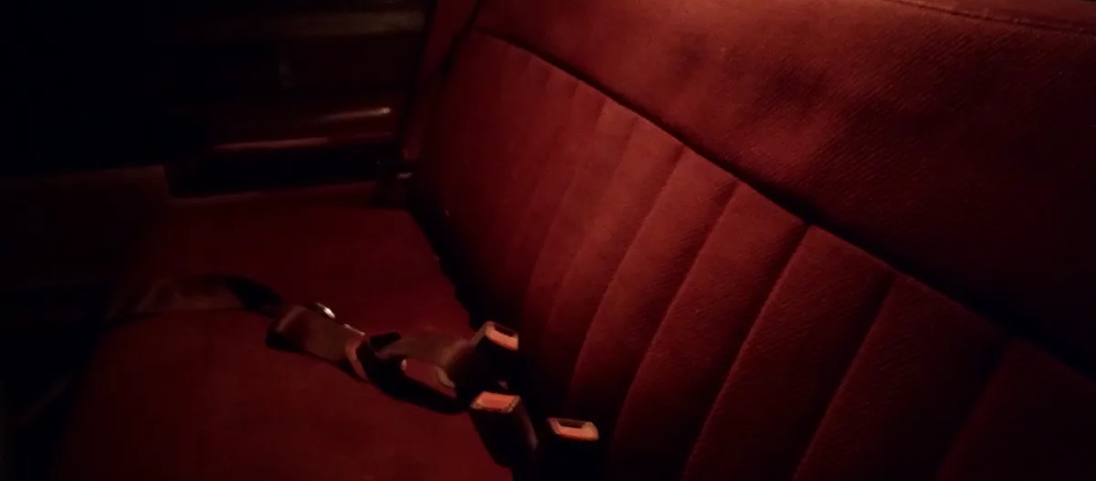 A dimly lit, close-up shot of a red car seat with a seatbelt buckle and strap lying loosely across the cushion. The image…