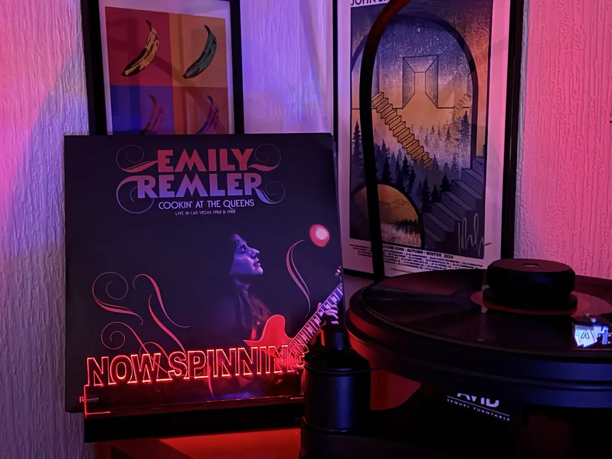 A vinyl record player with a record titled 'Emily Remler - Cookin' at the Queens' is shown next to a neon sign that reads…