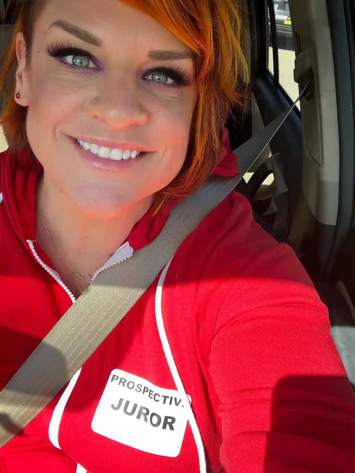 A woman with orange hair and a bright red hoodie is taking a selfie while sitting in a car, wearing a seatbelt. A white name…