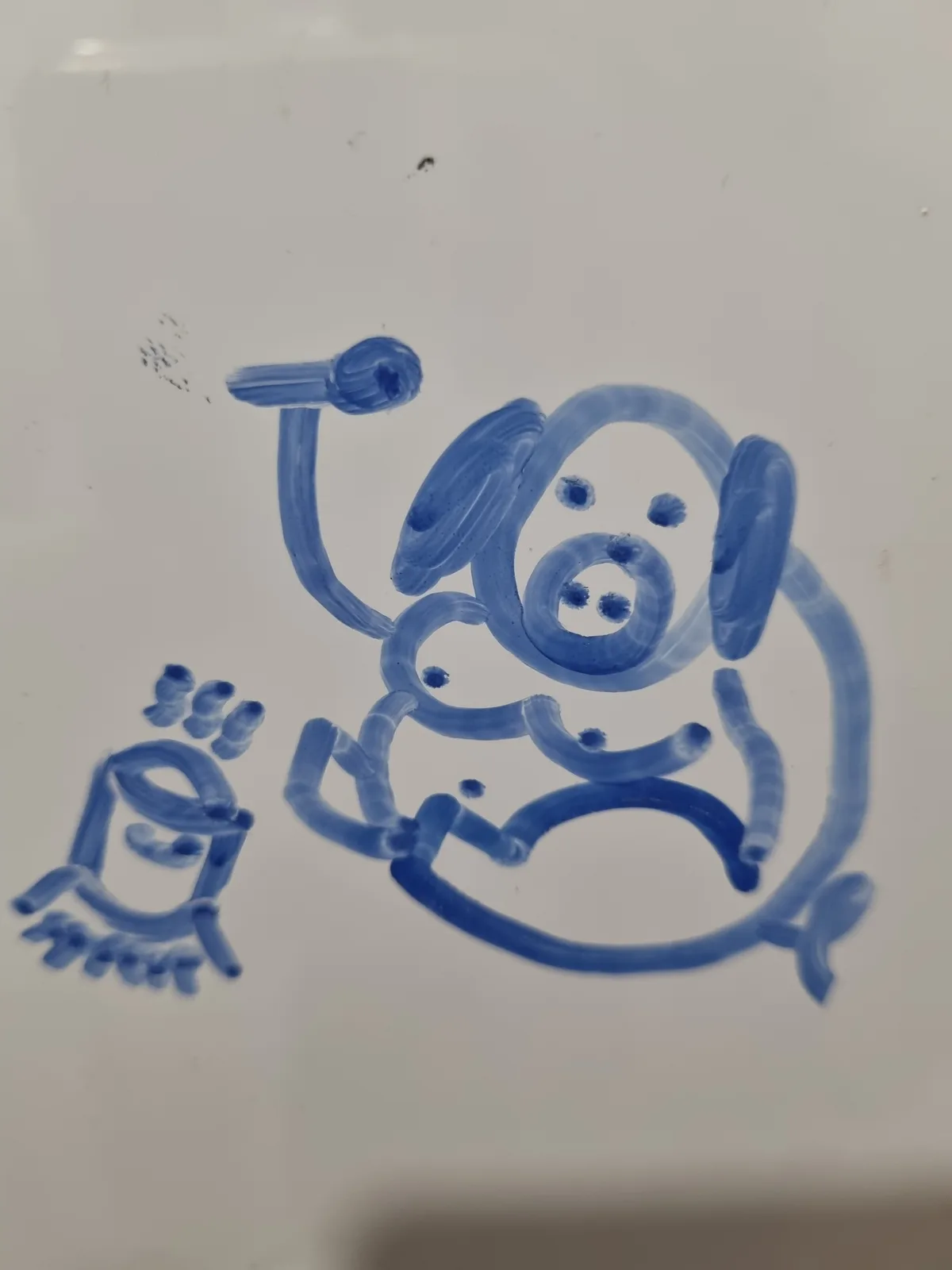 A child's blue crayon drawing on white paper shows a round, smiling creature with floppy ears, a snout, and a raised arm…