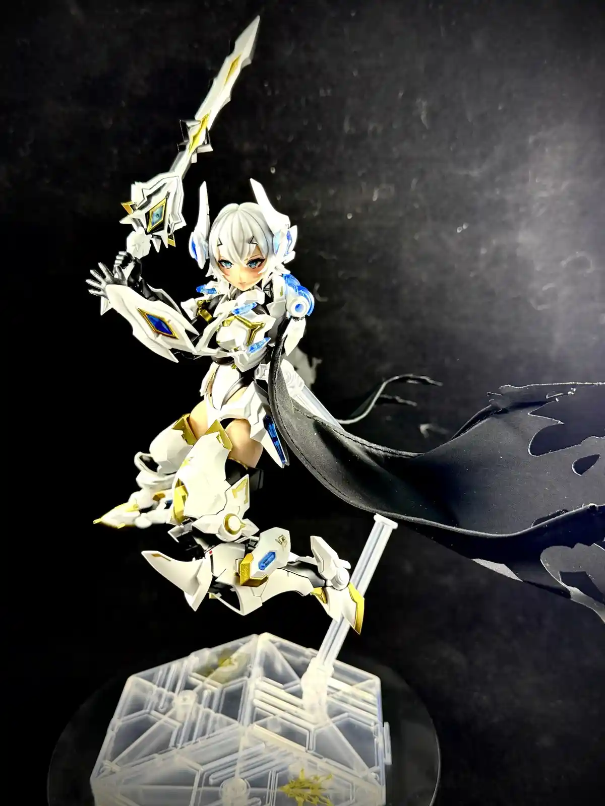A white and gold armored anime-style figurine with blue accents, holding a large sword and a black cape, posed dynamically…
